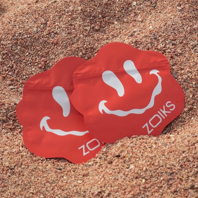 Zwei rote, blumenförmige Päckchen mit weißen, lächelnden Gesichtern und dem Wort „ZOIKS“ sind teilweise in grobem, rötlichem Sand vergraben – eine verspielte Szene, die selbst einen von Reiseangst geprägten Moment aufhellt.