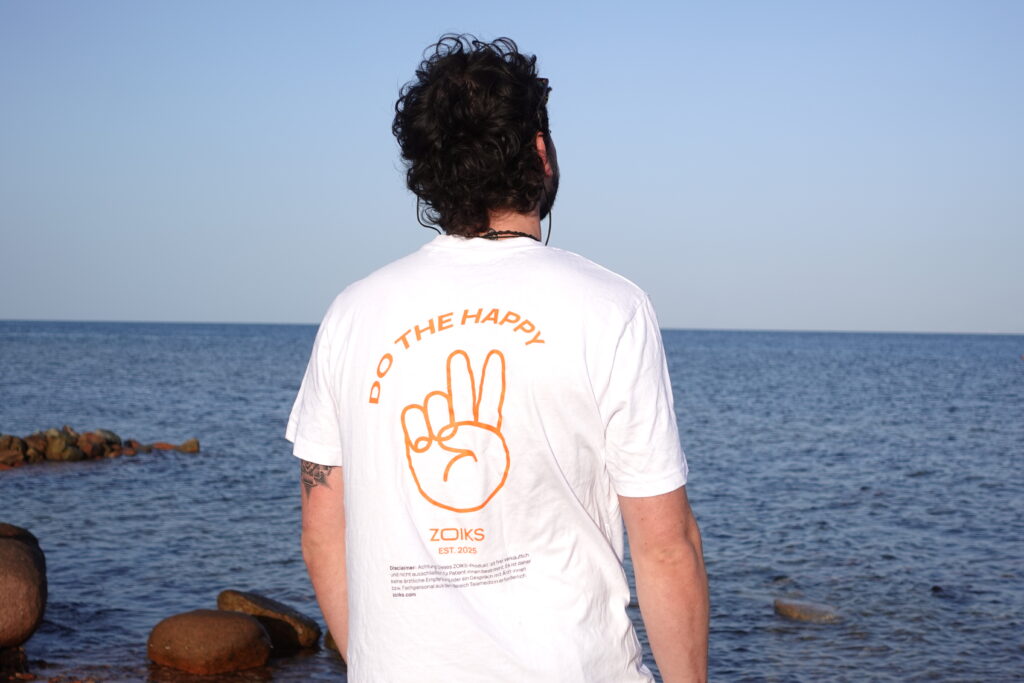 Eine Person mit lockigem Haar steht an einem felsigen Ufer mit Blick auf das Meer und trägt ein weißes Shirt, auf dessen Rückseite „DO THE HAPPY“ und ein orangefarbenes Handzeichen aufgedruckt sind. Trotz gelegentlicher Reiseangst steht sie unter einem strahlend blauen Himmel und genießt die Ruhe des weiten Horizonts.