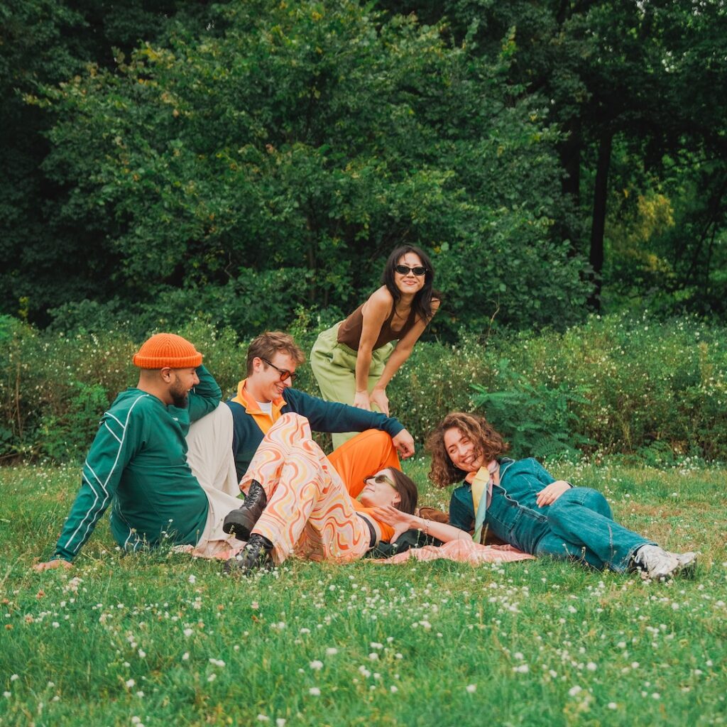 Fünf Freunde entspannen sich auf dem grünen Rasen eines Parks und lachen gemeinsam. Drei sitzen oder liegen, zwei lehnen sich von hinten heran. Die lebhafte Kulisse – mit farbenfroher Freizeitkleidung und den üppigen Bäumen und dem Grün rundherum – schafft eine fröhliche Szene.