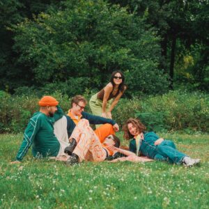 Fünf Freunde entspannen sich auf dem grünen Rasen eines Parks und lachen gemeinsam. Drei sitzen oder liegen, zwei lehnen sich von hinten heran. Die lebhafte Kulisse – mit farbenfroher Freizeitkleidung und den üppigen Bäumen und dem Grün rundherum – schafft eine fröhliche Szene.